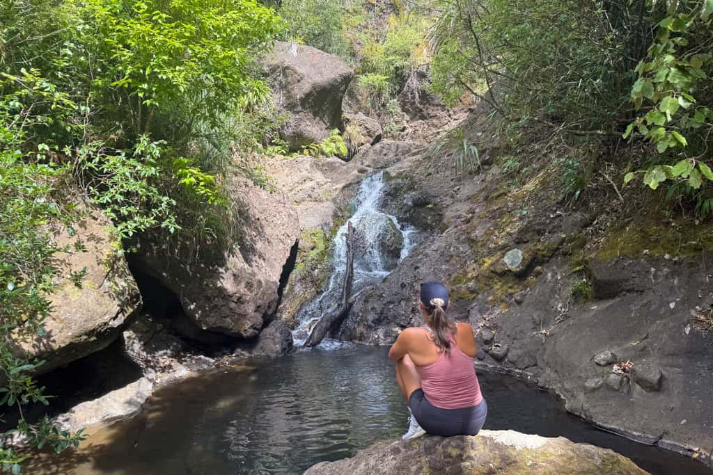 Woman sitting in front of a small waterfall on a rock.