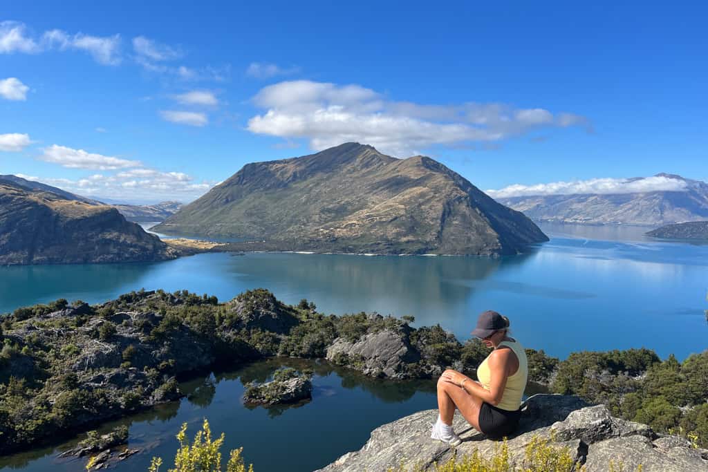 Woman sitting on the edge of a rock with a lake and mountains in the background.