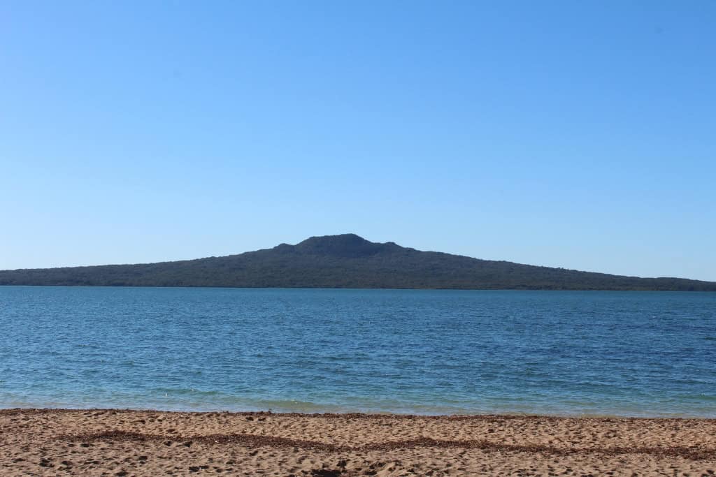 A view of Rangitoto Island from a sandy beach over the water.