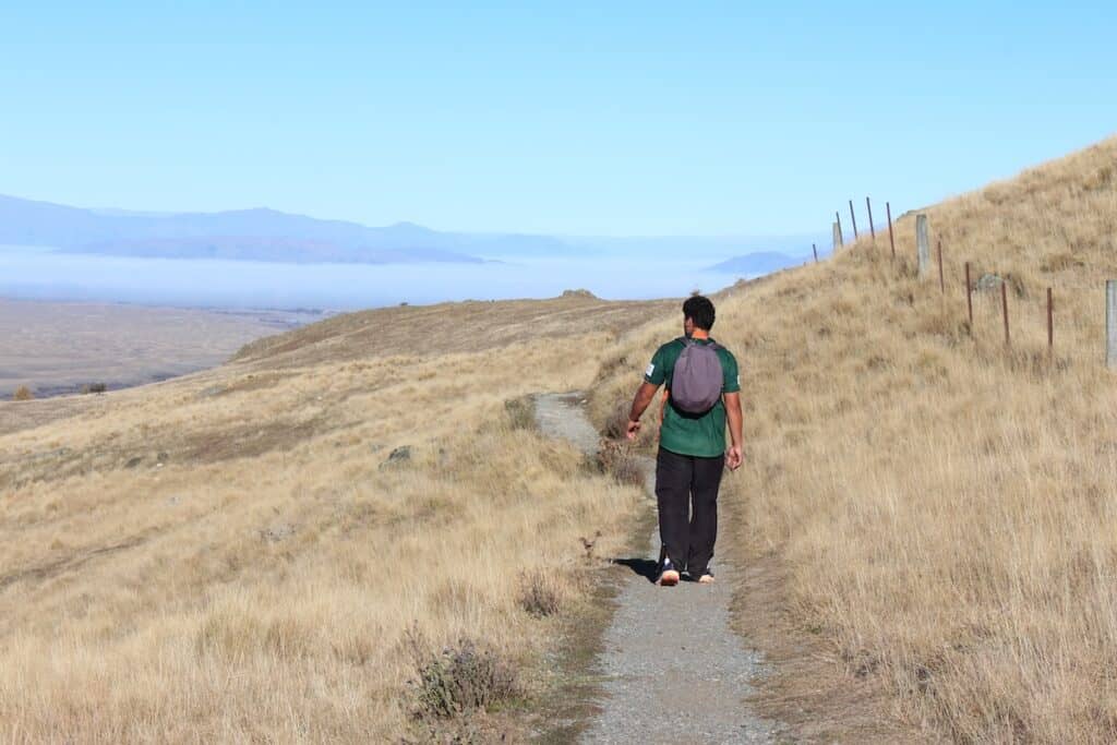 Man walking along a gravel track surrounded by grassy hills.