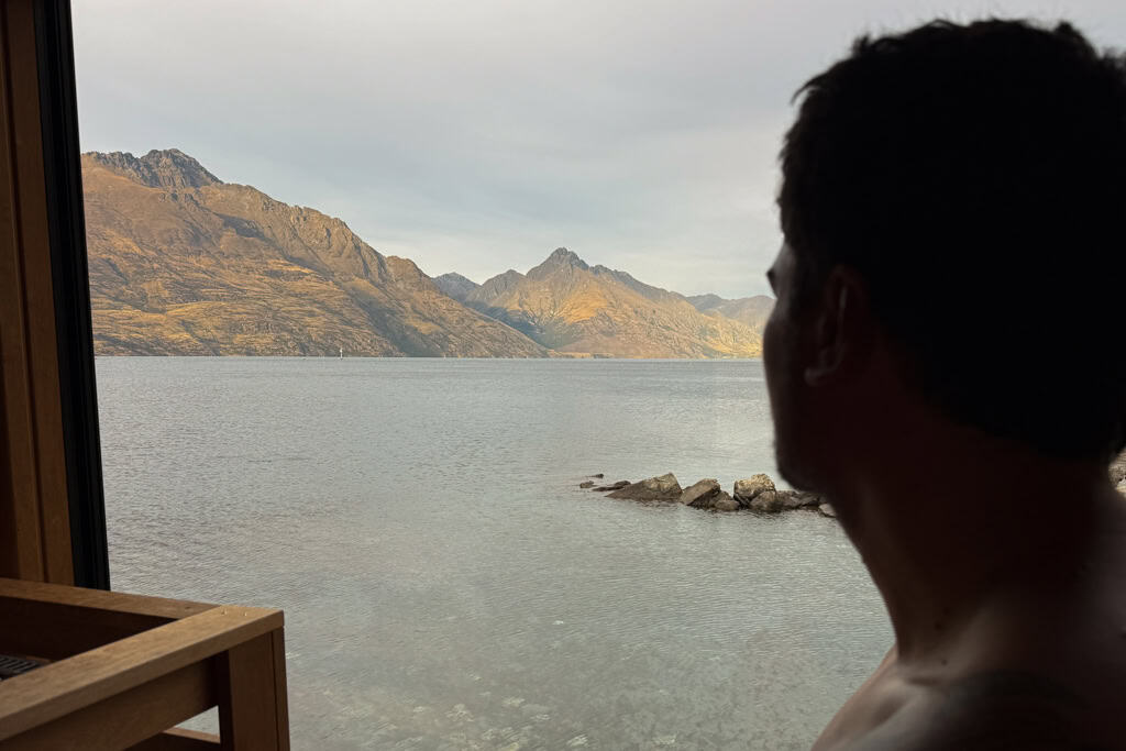 Man sitting in a sauna looking out over the lake at the mountains in the distance.