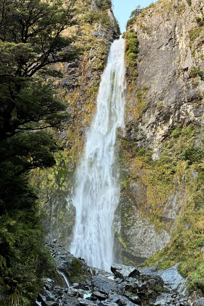 Devil’s Punchbowl Waterfall plunges down a tall, rugged rock face surrounded by dense native forest in Arthur’s Pass, New Zealand.